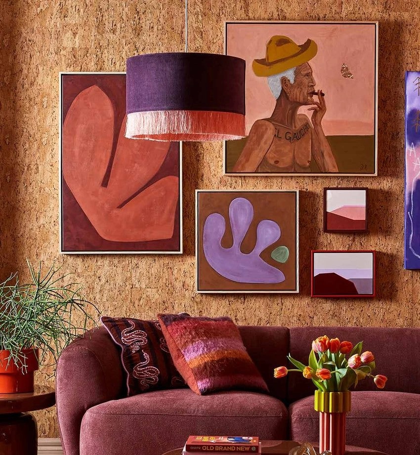 A living room with a purple sofa, a large fringed pendant lamp, and a gallery wall of abstract and figurative paintings on a cork-like wall.