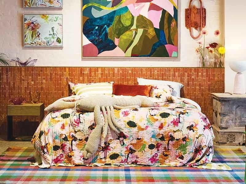 A colorful bedroom with a bed covered in a floral quilt, a striped rug, a textured headboard, and several abstract paintings on the wall.