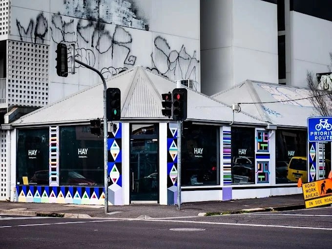 A street-level view of the HAY design store in Melbourne, located on a corner with traffic lights and a road work sign nearby.