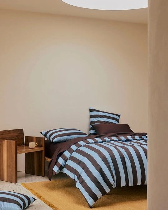 A bed with blue and brown striped bedding and a brown fitted sheet, next to a wooden side table with a white mug.