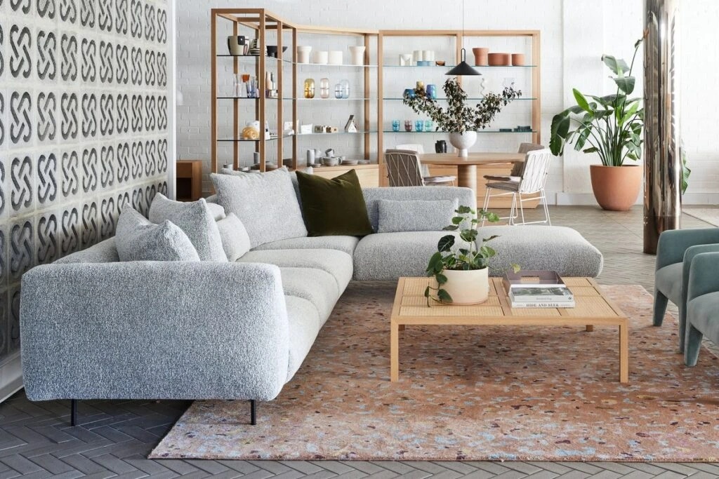 A cozy living room with a light-colored sectional sofa, a wooden coffee table, and a large shelving unit in the background.