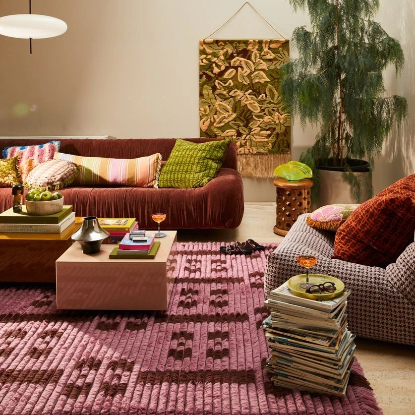 A cozy, bohemian-style living room featuring a brown velvet sofa, a pink tufted rug, and a variety of colorful pillows and decor.