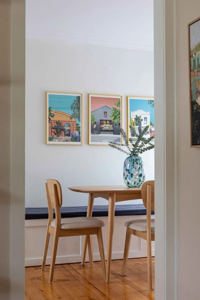 A cozy dining area featuring a light wood table, chairs, and a bench with three colorful art prints on the wall.