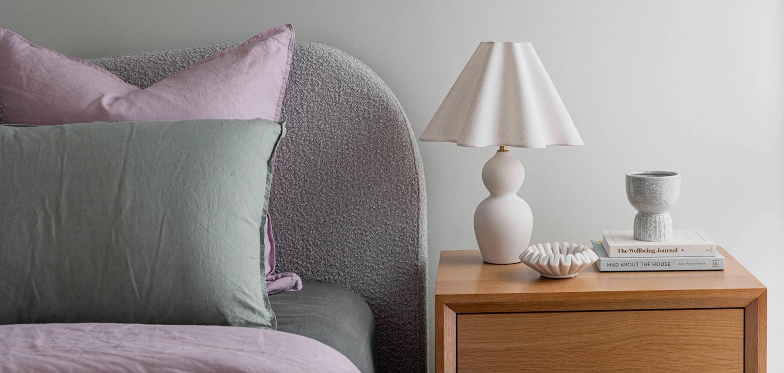 A close-up photo of a bed with a textured headboard and a wooden nightstand with a lamp, books, and decorative objects.