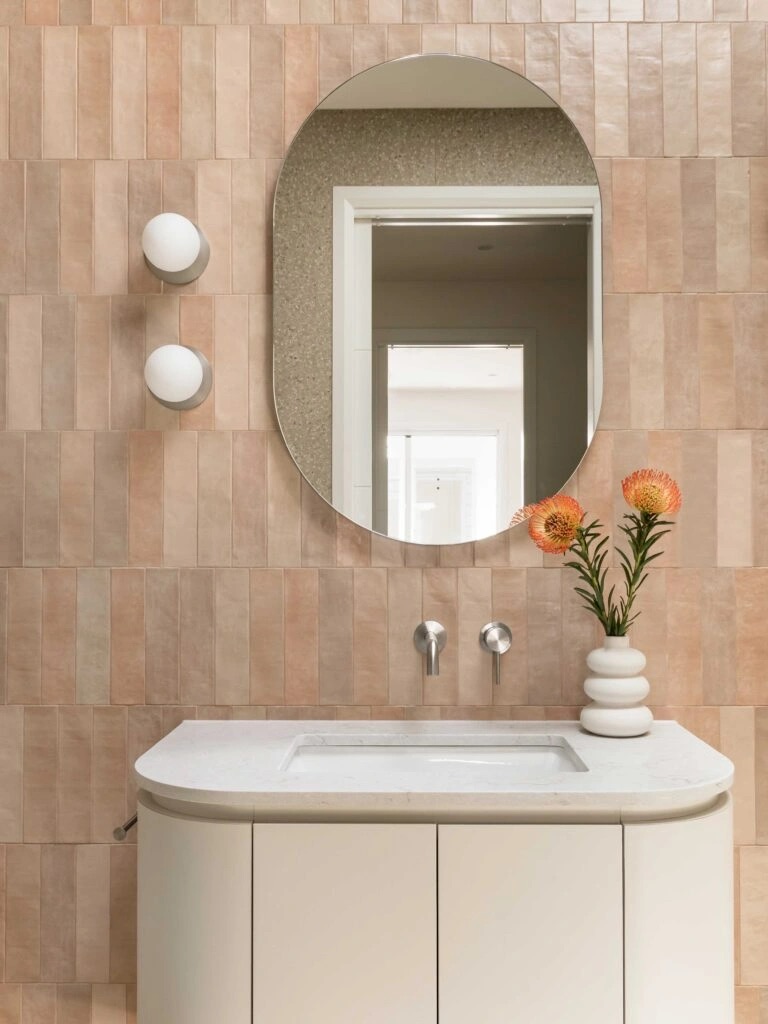 Photo of a white bathroom countertop and sink, an oval mirror, and vertical beige tiles on the wall.