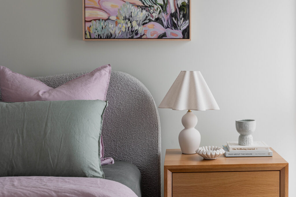 Interior decoration guide showing styled bedroom with curved upholstered bedhead, layered cushions, timber bedside table and ceramic lamp